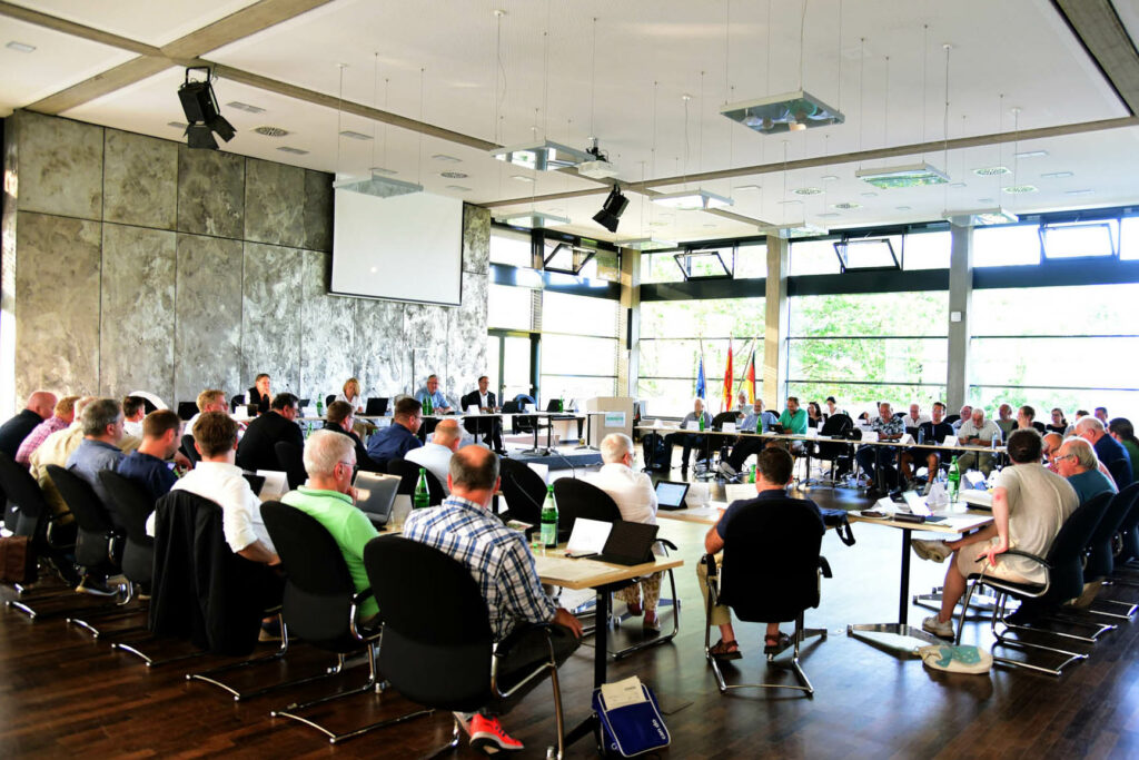 Eine Gruppe von Personen, die in einem modernen Konferenzraum mit großen Fenstern sitzen und an einer formellen Sitzung oder Diskussion teilnehmen.