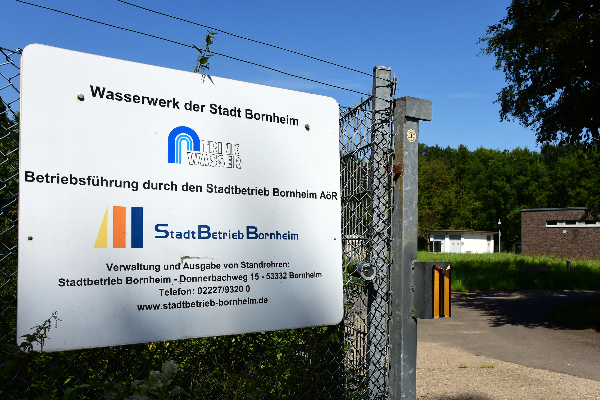 Schild am Eingang des Wasserwerks Bornheim mit Angaben zur Verwaltung und zu den Ansprechpartnern in deutscher Sprache, im Hintergrund ein Gebäude und eine Grünanlage.