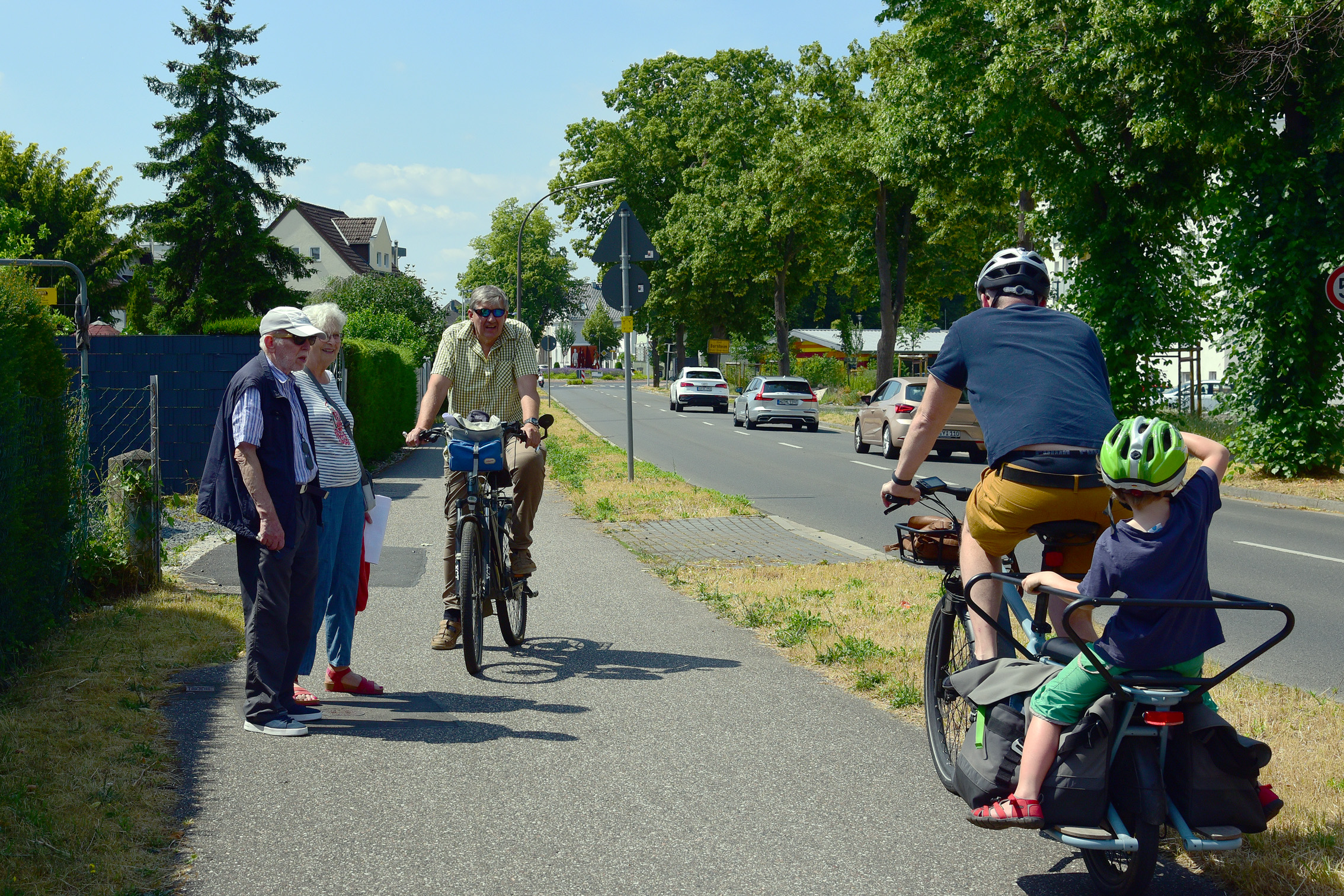 Zwei Radfahrer und zwei Fußgänger teilen sich einen Gehweg in der Nähe einer Straße mit vorbeifahrenden Autos; ein Radfahrer trägt ein Kind mit einem Helm auf dem Rücksitz.