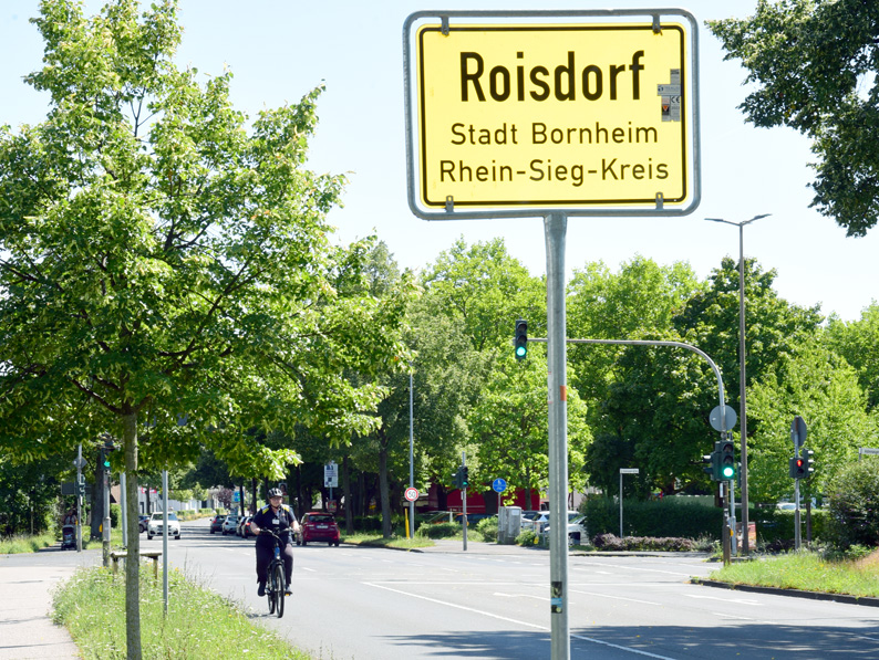 Eine Straßenszene mit einem Radfahrer und einem gelben Ortsschild mit der Aufschrift "Roisdorf, Stadt Bornheim, Rhein-Sieg-Kreis", umgeben von Bäumen und Ampeln.