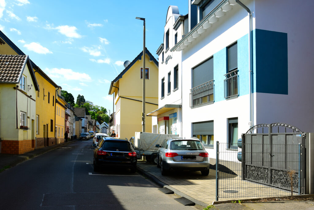 Eine schmale Wohnstraße mit geparkten Autos, modernen und älteren Häusern und einem strahlend blauen Himmel mit vereinzelten Wolken.
