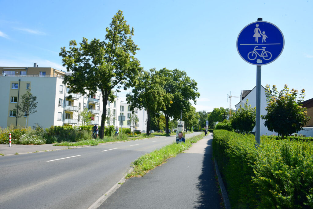 Eine Straße mit einem separaten Fußgänger- und Fahrradweg, gekennzeichnet durch ein blaues Schild, gesäumt von Bäumen und modernen Wohnhäusern an einem sonnigen Tag.