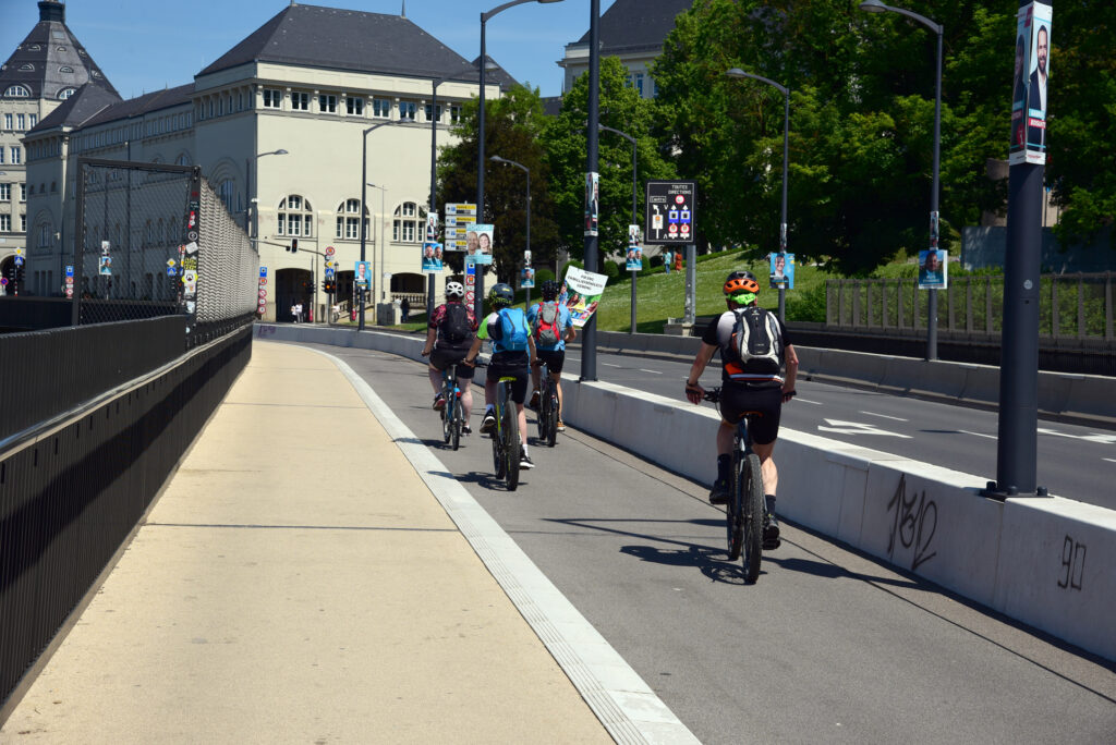 Vier Radfahrer fahren auf einer Stadtstraße in der Nähe eines großen Gebäudes, im Hintergrund sind Grünflächen und verschiedene Plakate an Lichtmasten zu sehen.
