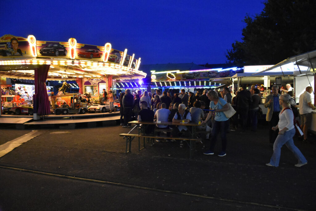 Die Menschen versammeln sich nachts auf einem Jahrmarkt, mit einem hell erleuchteten Karussell auf der linken Seite und Essensständen auf der rechten Seite.