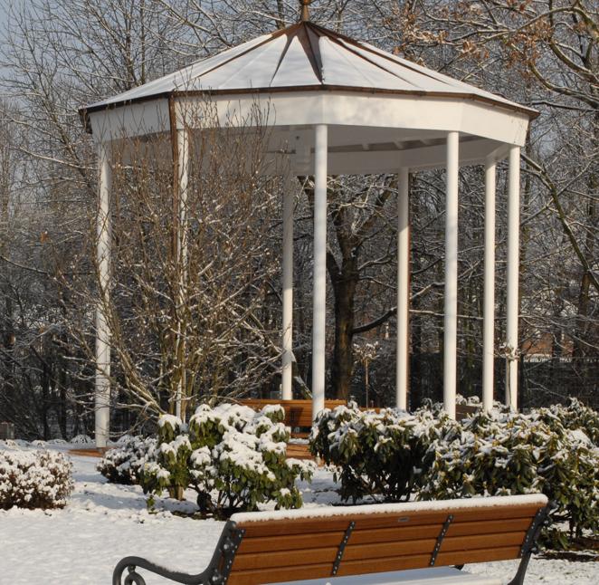 Ein weißer Pavillon mit Spitzdach steht in einem verschneiten Park, umgeben von blattlosen Bäumen und Sträuchern, mit einer Holzbank im Vordergrund.