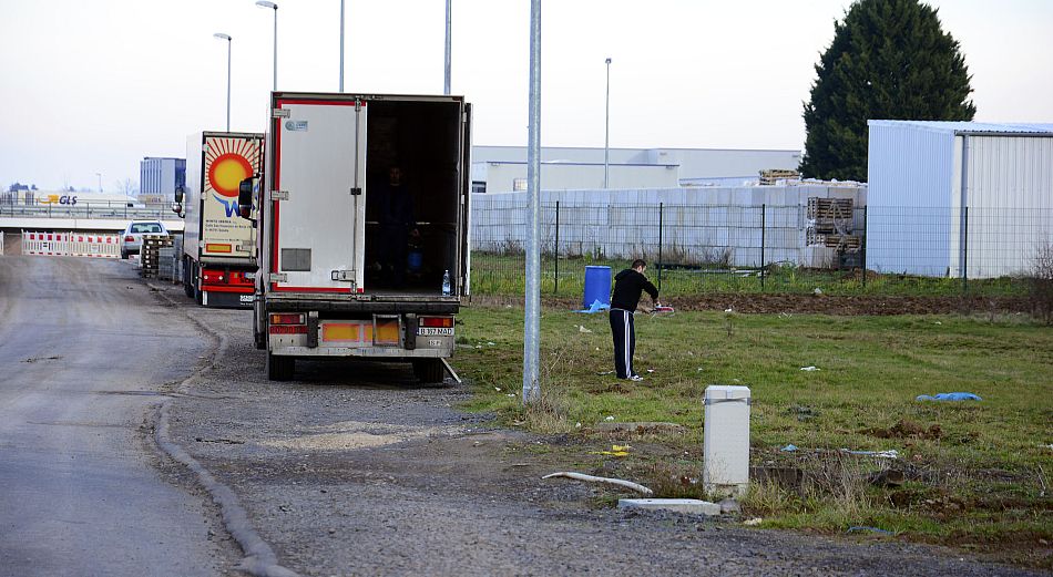 Eine Person steht auf einer Wiese in der Nähe einer Straße, während in der Nähe ein Lastwagen mit geöffneten Hintertüren geparkt ist; im Hintergrund sind Industriegebäude zu sehen.