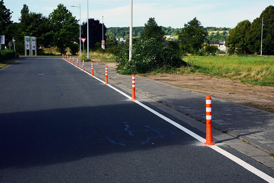 Orangefarbene und weiße Verkehrspoller säumen den Rand einer gepflasterten Straße neben einer Grünfläche und trennen die Straße vom Gehweg.