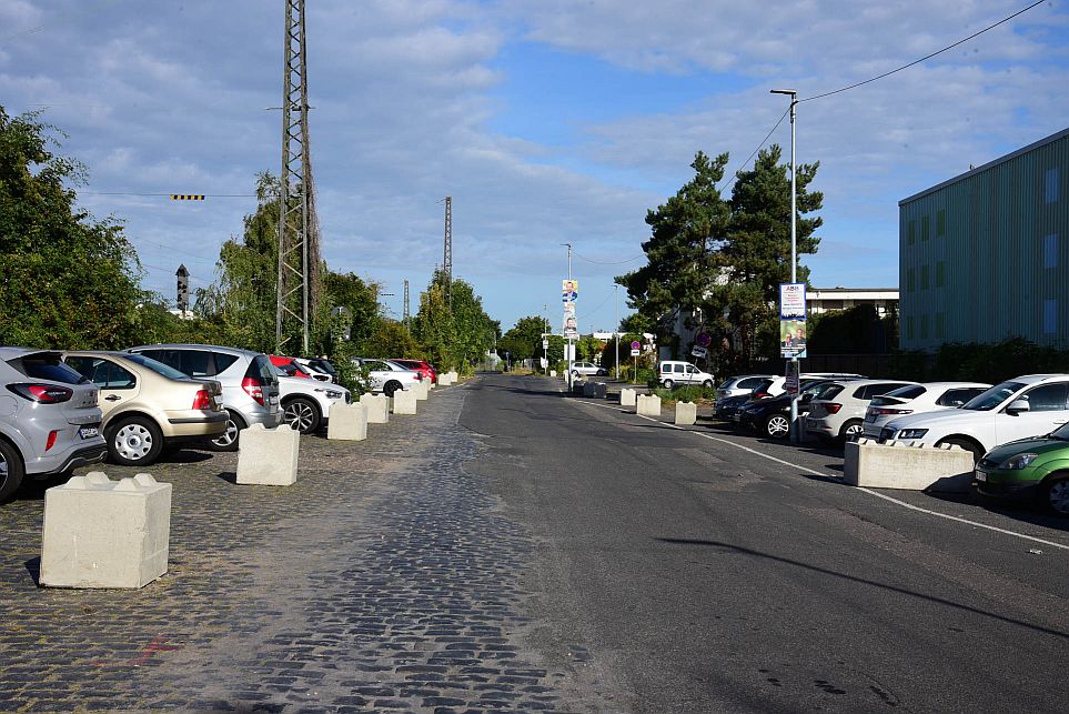 Eine Straße mit geparkten Autos auf beiden Seiten, gesäumt von Bäumen und Gebäuden, unter einem teilweise bewölkten Himmel. Betonbarrieren trennen den Parkbereich von der Straße.