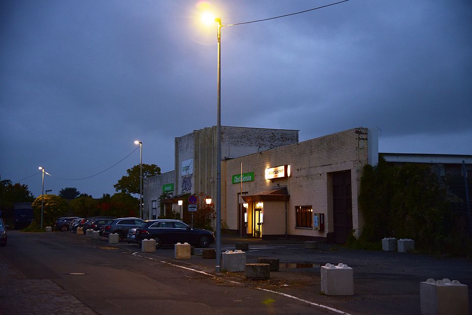 Eine schwach beleuchtete Straße am Abend mit geparkten Autos und einer Reihe weißer Backsteinhäuser unter einem bewölkten Himmel.