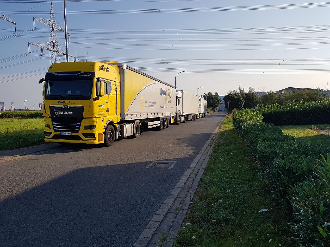 Ein gelber MAN-Sattelzug mit weißem Anhänger ist am Straßenrand geparkt, ein weiterer Anhänger ist dahinter zu sehen. Im Hintergrund sind Stromleitungen und Grünflächen zu sehen.