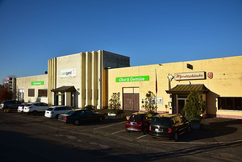 Ein gelbes Geschäftshaus mit den Schildern "Landgard", "Obst & Gemüse" und "Markthalle", davor geparkte Autos unter einem strahlend blauen Himmel.