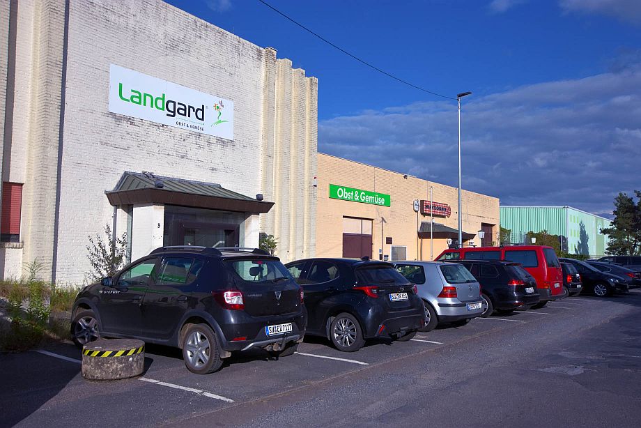 Eine Reihe geparkter Autos vor einem Industriegebäude mit den Schildern "Landgard" und "Obst & Gemüse" an den Außenwänden.