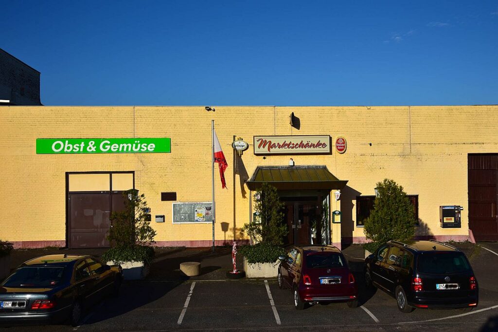 Ein gelbes Gebäude mit einem Schild "Obst & Gemüse" und einem weiteren Eingang mit der Aufschrift "Marktschänke", davor drei geparkte Autos und Topfpflanzen.
