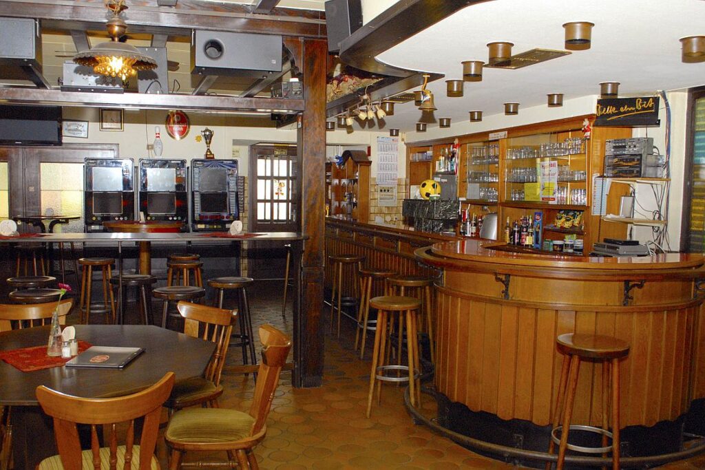 Eine leere Bar mit Holzhockern und -tischen, Glaswaren in Regalen hinter dem Tresen und Spielautomaten an der Rückwand.