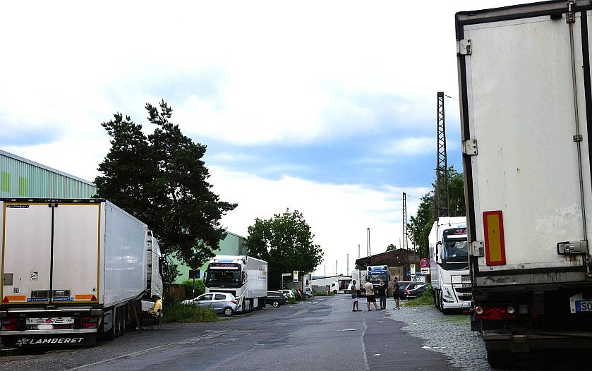 Mehrere geparkte Lastwagen säumen beide Seiten einer schmalen Industriestraße, ein paar Menschen gehen spazieren und Autos parken in der Nähe grüner Gebäude unter einem bewölkten Himmel.
