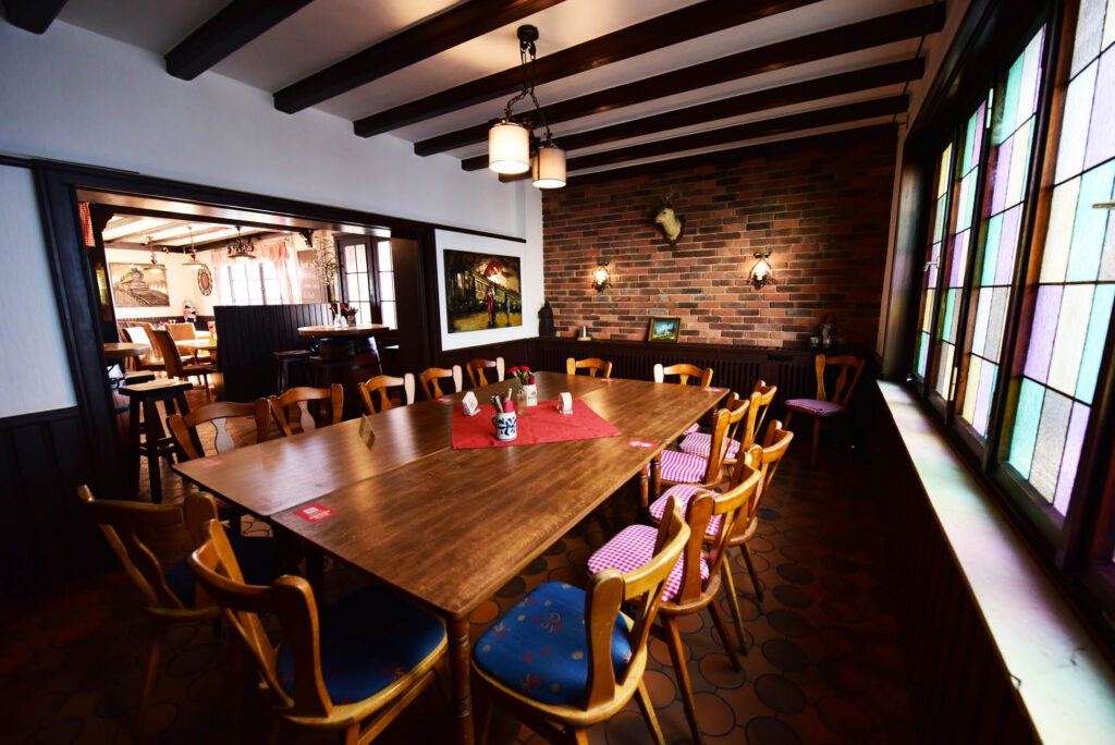 Ein hölzerner Esstisch mit Stühlen steht in einem rustikalen Raum mit Backsteinwänden, Holzbalken an der Decke und Buntglasfenstern.