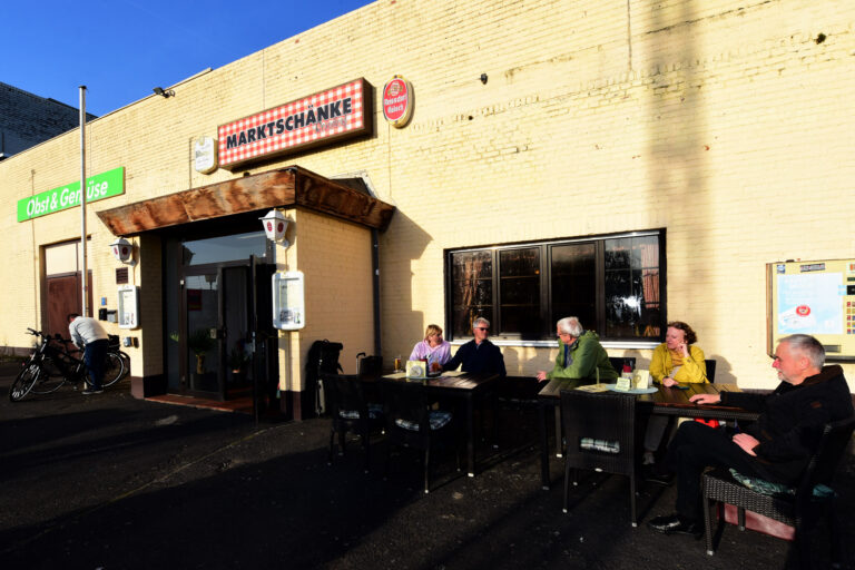 Menschen sitzen an Tischen im Freien vor einem beigen Gebäude mit dem Schild "Marktschänke"; eine Person steht neben einem Fahrrad am Eingang.