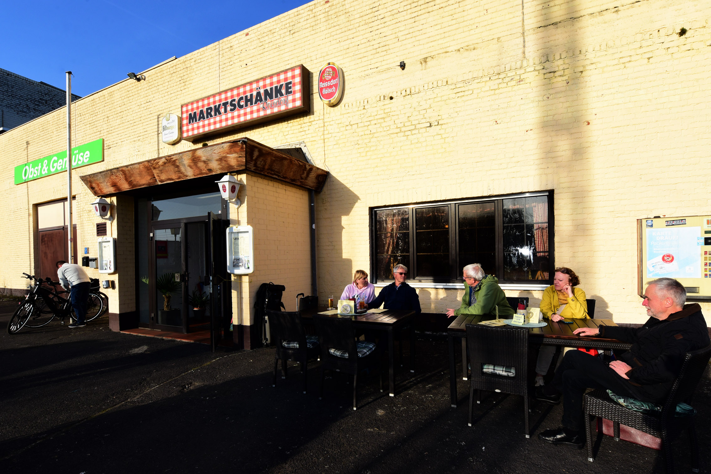 Menschen sitzen an Tischen im Freien vor einem beigen Gebäude mit dem Schild "Marktschänke"; eine Person steht neben einem Fahrrad am Eingang.