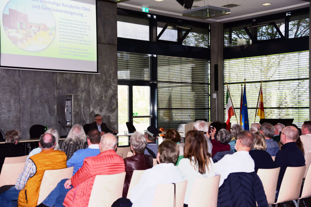 Eine Gruppe von Personen sitzt in einem Konferenzraum und hört einem Vortragenden zu, dessen Präsentation auf eine Leinwand projiziert wird und dessen Fahnen an den Fenstern zu sehen sind.