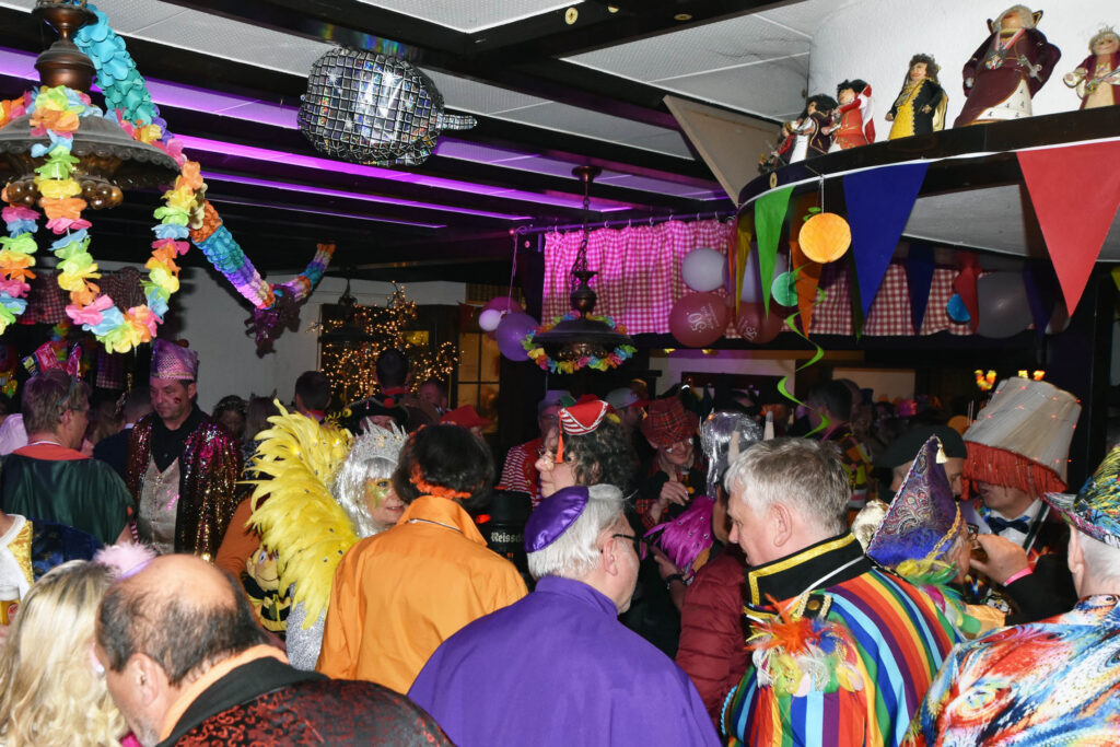 Eine überfüllte Indoor-Party mit Menschen in bunten Kostümen und Hüten, festlicher Dekoration wie Luftschlangen, Luftballons, Fahnen und einer Discokugel über dem Kopf.