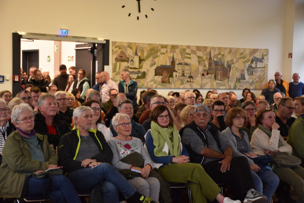 Eine große Gruppe Erwachsener sitzt eng beieinander in einem Raum und blickt aufmerksam nach vorn. Im Hintergrund sind eine Wanduhr und eine Wandmalerei zu sehen.