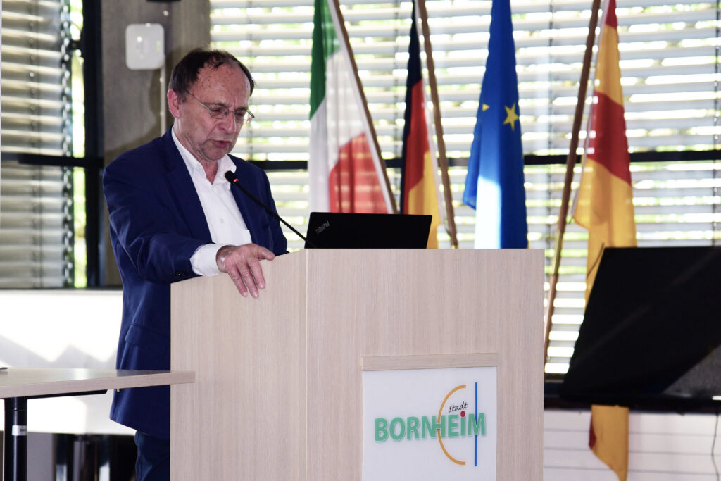 Ein Mann in einem blauen Anzug spricht an einem Podium mit einem Laptop; im Hintergrund sind mehrere Fahnen zu sehen. Das Podium ist mit "Stadt Bornheim" beschriftet.