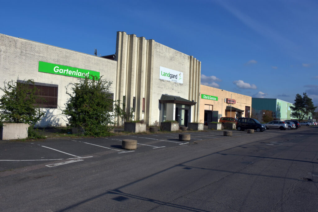Eine Reihe von Geschäftshäusern mit Schildern für Gartenland, Landgard und Oase Gartenwelt; davor ein meist leerer Parkplatz unter freiem Himmel.