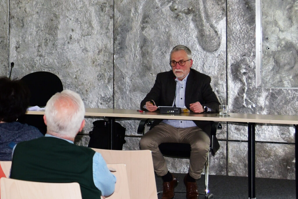 Ein älterer Mann im Blazer spricht an einem Tisch vor einem Publikum, mit einer grauen Steinmauer als Hintergrund.