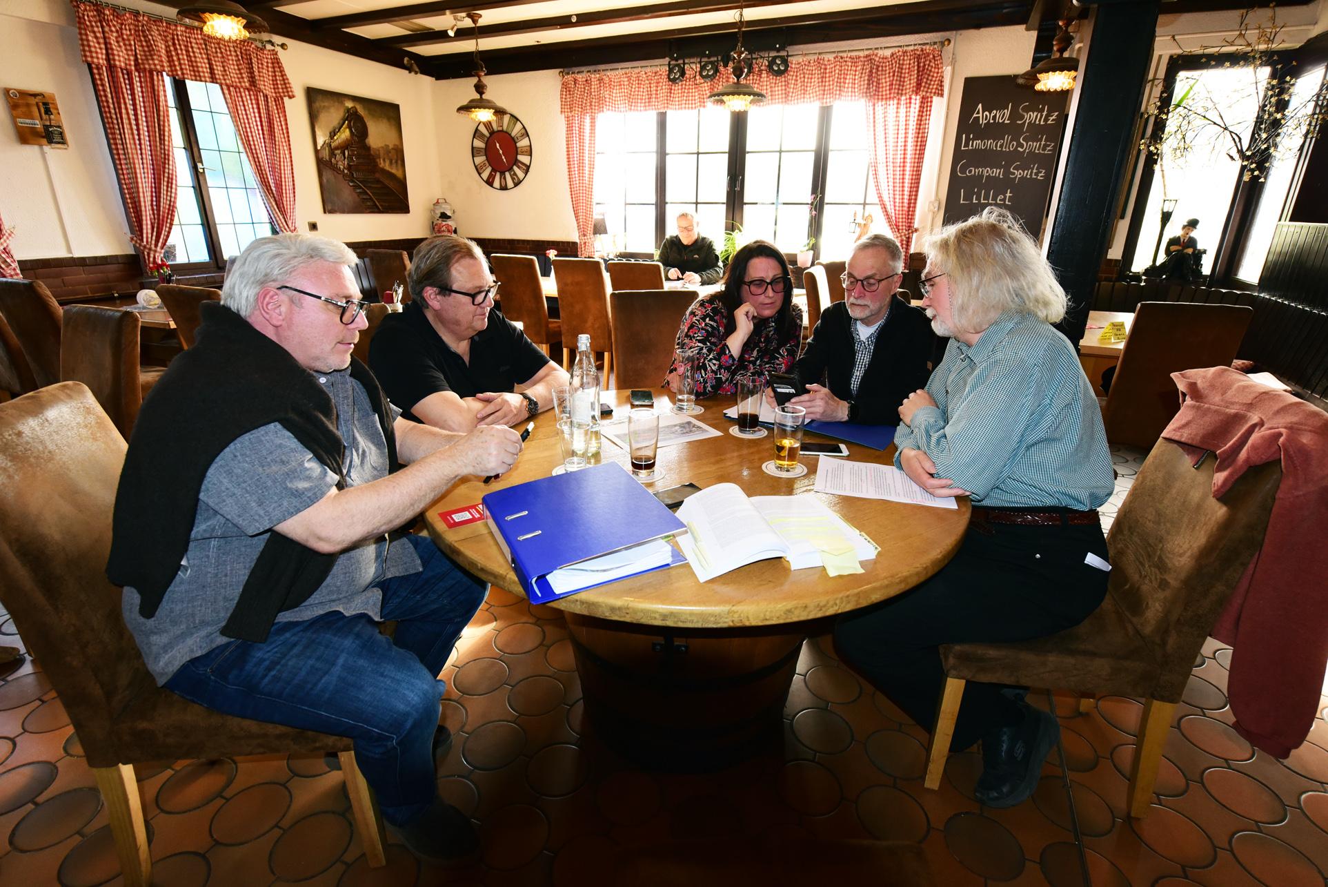 Fünf Erwachsene sitzen in einem Restaurant um einen runden Holztisch herum und unterhalten sich, während auf dem Tisch Unterlagen, Gläser und Getränke zu sehen sind.